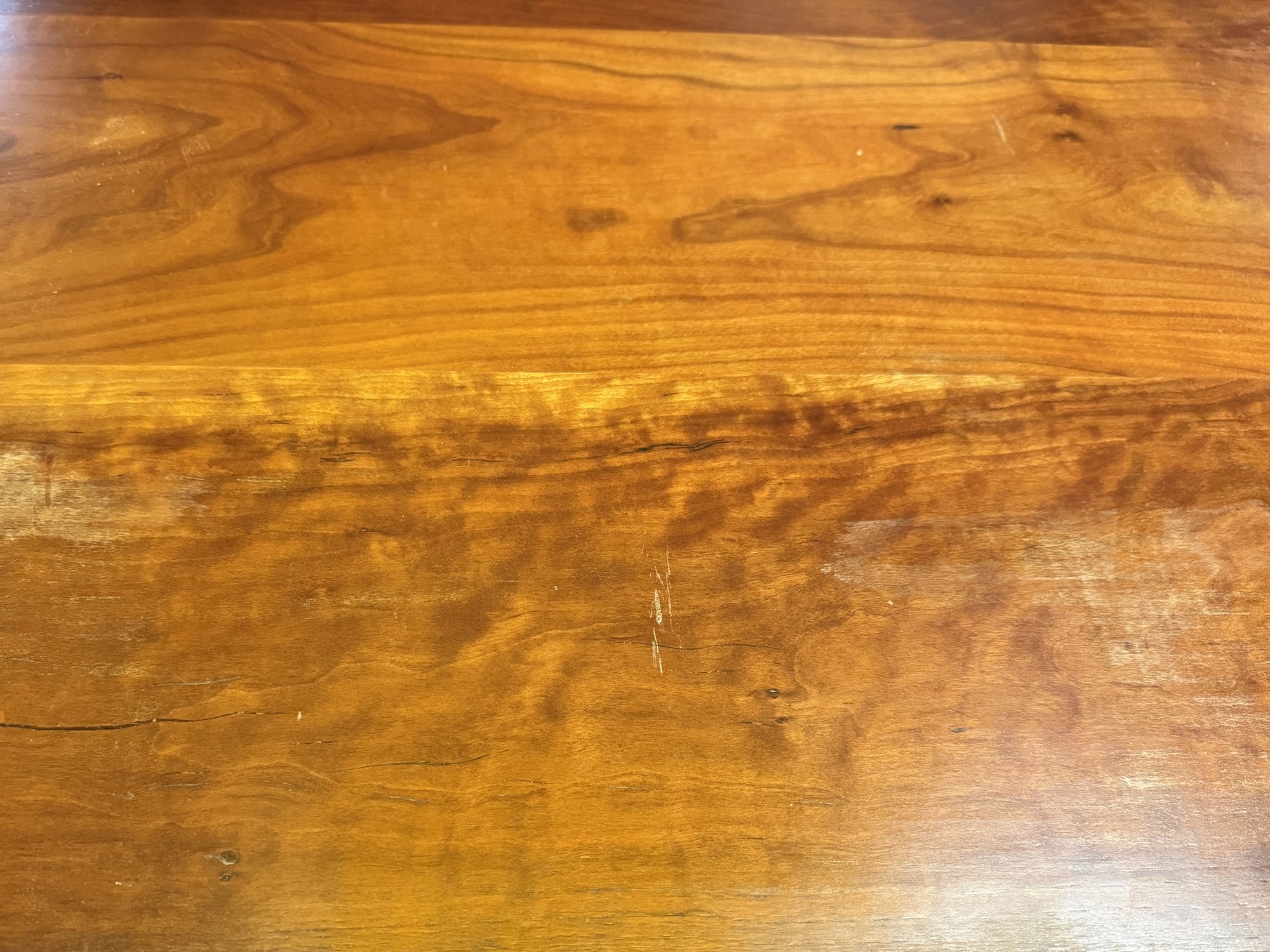 Section of a hardwood dining tabletop before refinishing, with white worn patches and scratches in the old finish